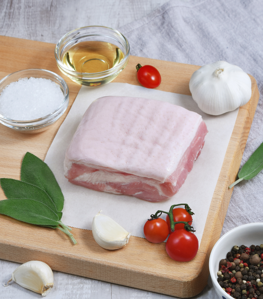 Large serving of fresh and clean Wicks Manor English Pork Belly on a wooden board with garlic, red grape tomato and spices by side